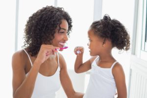 A woman and little girl facing each other smiling and brushing their teeth