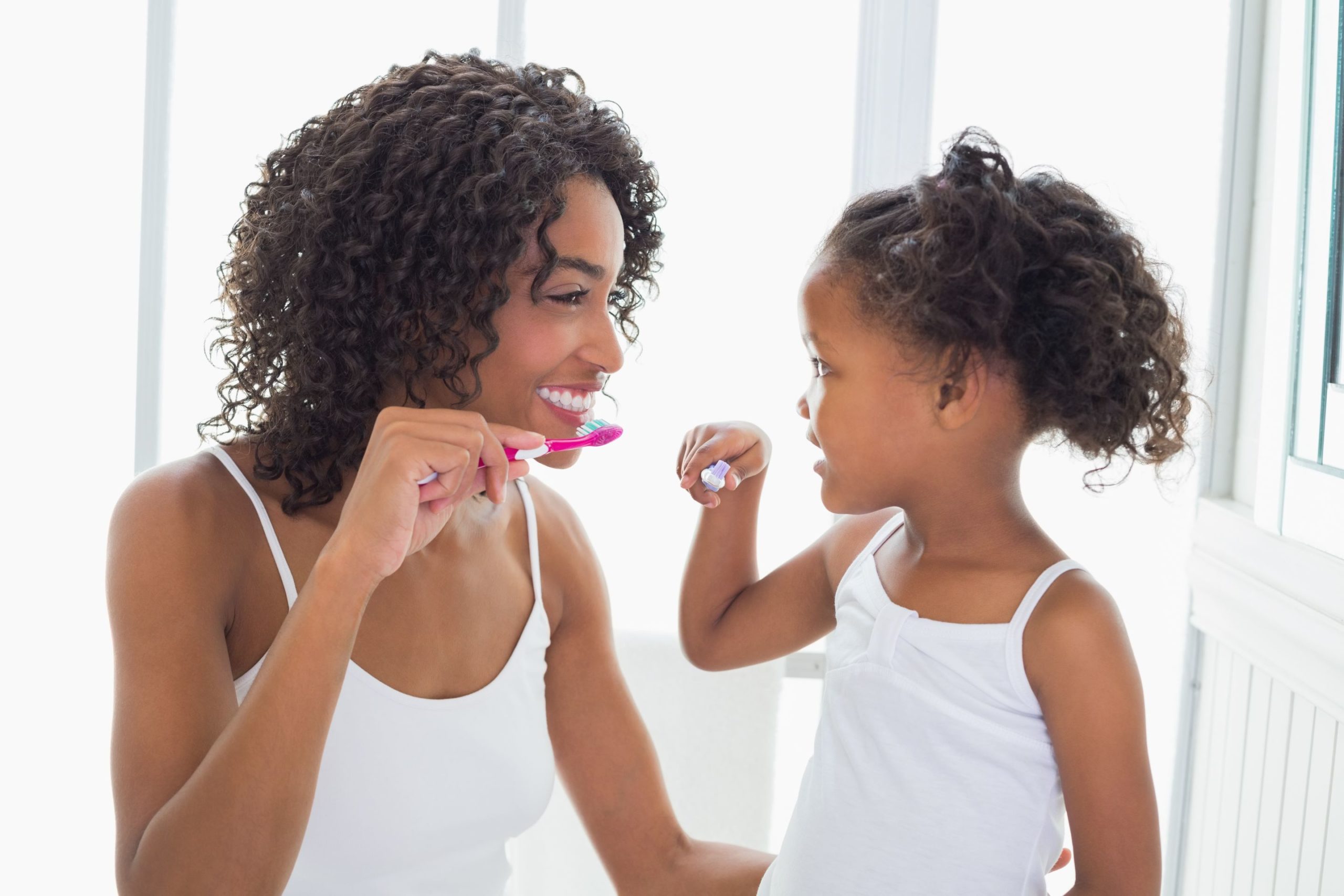 A woman and little girl facing each other smiling and brushing their teeth.