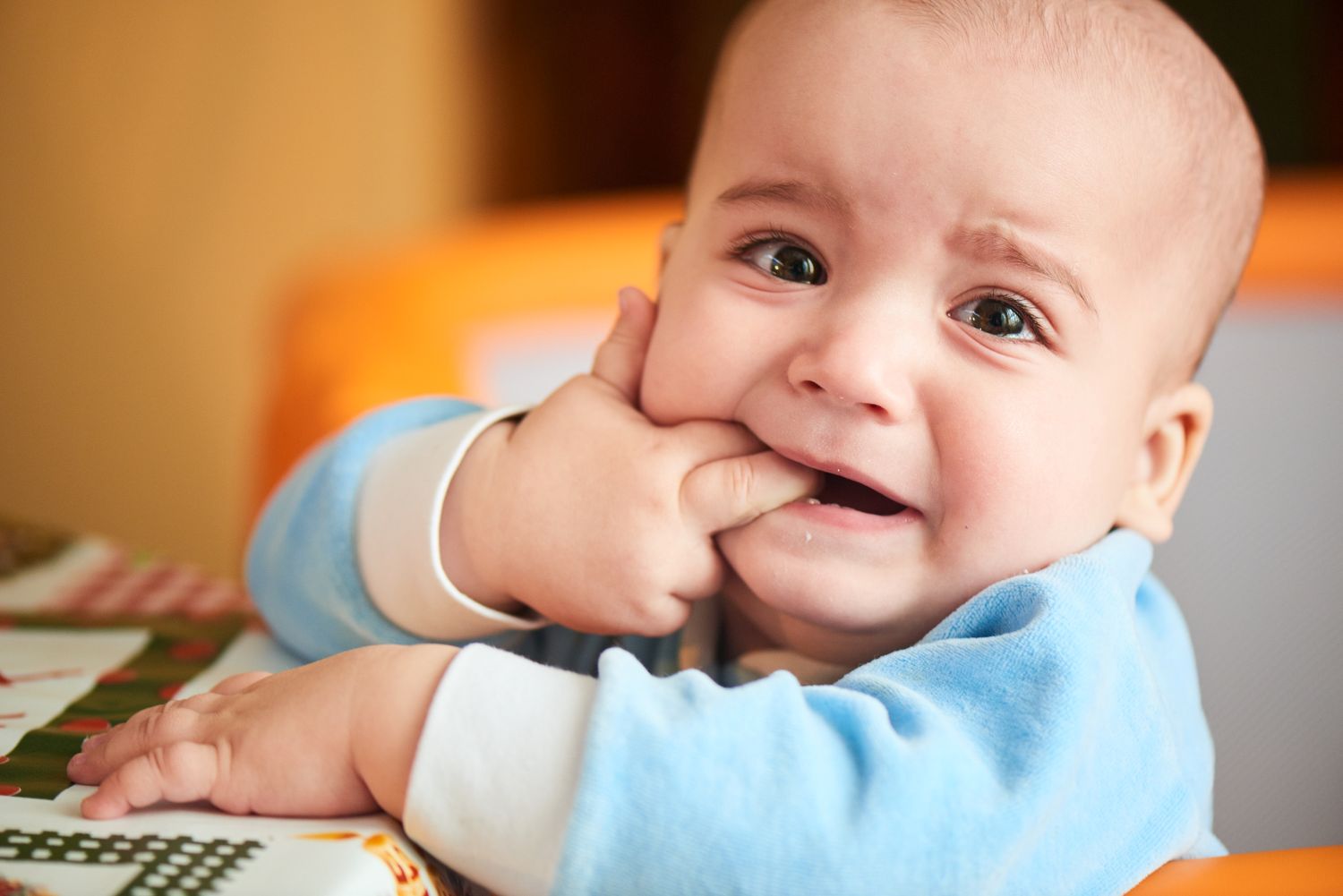 Infant in blue onesie chewing fingers with tears in eyes.